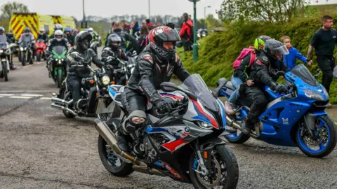 Dozens of people on motorbikes line up on a road. They are dressed in black and wearing black helmets. 