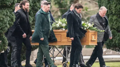 PA Media A light brown wooden coffin adorned with white flowers is accompanied by mourners.