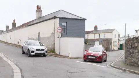 James Road and Le Mont Pinel intersection in a residential area, with two cars approaching from opposite directions — a white SUV on the left side of the road and a red car on the right. The buildings lining the street are mostly light‑coloured, with one grey‑panelled upper floor on the corner. A stone wall borders the right side of the road, and the sky is overcast. A “no entry” traffic sign is visible on the corner building.