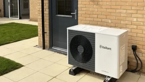 A white heat pump outside a house. It is to the right of the front door.