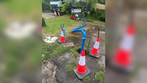 Stella Spiteri A grassed garden with a blue hose pipe entering the ground, surrounded by three orange cones. 