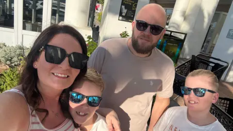 A sunny outdoor selfie shows four people wearing sunglasses gathered close together outside a modern building with white columns and large windows.
Two adults and two children are visible in the foreground, dressed casually in light-coloured summer clothing, with landscaped plants and paved walkways surrounding them.
