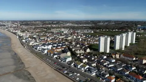 BBC An aerial photograph of Jersey's east coast in St Clement. It is a highly residential area, with several homes and four white tower blocks. A two-lane road is along the coast. The beach is on the left of the image, with the tide quite high up the bay.