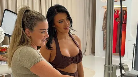 MURCI The two women in a studio, stood looking at a laptop screen and smiling. Meagan has long brown hair and wears a brown crop top, and Olivia's blonde hair is tied back, she is wearing a grey top. 