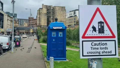 @ASignOfHumour A photo of an old-fashioned blue police box. It looks like a telephone box but was used by the police in years gone by. The sign has a drawing of the Doctor Who tardis, which is a police box, and the doctor nearby. It says Caution: Time Lords crossing ahead.