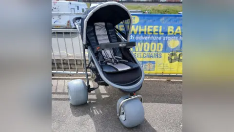 A wheelchair buggy is in a car park. It looks like a sturdy pushchair with large inflatable grey wheels.