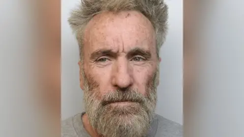 Police custody image of Tony Devenport with dishevelled grey hair and beard wearing a grey top. 