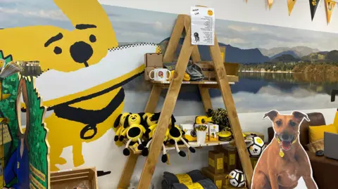 The walls of the dog hub are decorated with paintings of dogs and local landscapes. A cartoon cut out of a dog is placed next to a shelf with plush toys and mugs.
