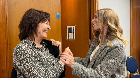 Harriet Heywood/BBC Ali Harris meeting her Petals counsellor, Sue Brookes. The pair are greeting each other warmly, smiling and holding hands in a quiet moment of reconnection. They are indoors, in front of a wooden door marked with a blue sign. They are both smiling widely as they take each other in. 