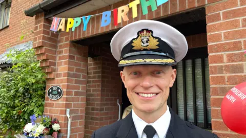 A smiling man in uniform with a banner saying Happy Birthday behind him