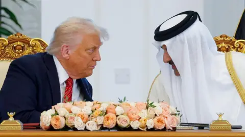 Trump and Al Thani lean towards each other in conversation behind an ornate gold desk with a bunch of white and orange and pink flowers on it 