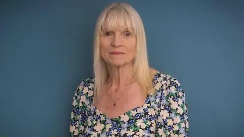 Angharad Stone Dr Denise Chaffer - a middle-aged woman with light blonde, shoulder-length hair. She is looking directly at the camera and is wearing a blue, green and cream floral print sleeved dress and is wearing a necklace with a small dark pendant on it. Denise has a neutral expression on her face and is standing in front of an all-blue backdrop