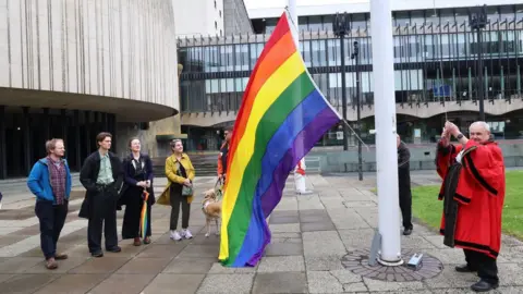 Newcastle City Council Lord Mayor Henry Gallagher, dressed in his red ceremonial robes, hoists a large Pride flag on a pole outside the Civic Centre. A group of people watch the ceremony.