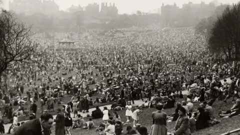 A black-and-white photograph, taken from the top of a hill, shows large crowds of people. They are at an outdoor event, with the silhouette of buildings along the skyline in the background.
