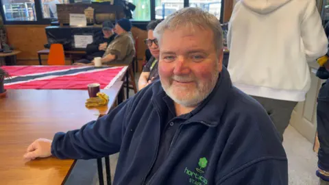 A man in a Horticap staff jacket sits at a table with gloves, a mug, and a red‑and‑black flag, while others chat in the background.