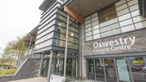 A large building with lots of windows and a stone clad front with the words Oswestry Leisure Centre on the front