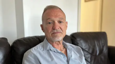David Thompson is a man with grey hair and a white goatee beard. He is wearing a pale blue, grey shirt with a silver chain around his neck. He is sitting on a dark brown leather sofa in front of a white wall, looking into the camera.
