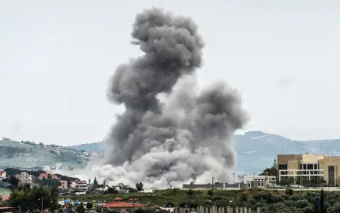 Abbas Fakih / AFP via Getty Images A large grey bundle of smoke rising from a village on a hillside. The smoke cloud fills most of the picture and is dramatic against the backdrop with village buildings in the foreground and hills in the background.