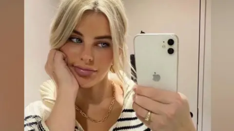 The photo shows Paris Wilson taking a mirror selfie indoors. They are holding a white iPhone with a dual‑camera setup in their right hand, and they are wearing a chunky striped knit top with a zip collar. Their hair appears to be light blonde and tied back loosely. A gold chain necklace is visible around their neck, and they are wearing a gold ring on their right hand. The setting looks like a simple, neutral-toned room, possibly a bedroom or changing area.