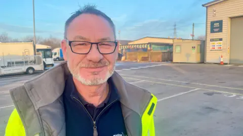 A man in a hi-vis jacket at a depot. There are vehicles and bins in the background. He has a beard and glasses and he is smiling. It is a bright and clear day with a blue sky.