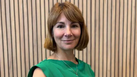 Lisa Spivey, a middle aged woman with a honey brown bob wearing a sleeveless green dress, stands against wooden panelling. She is smiling.