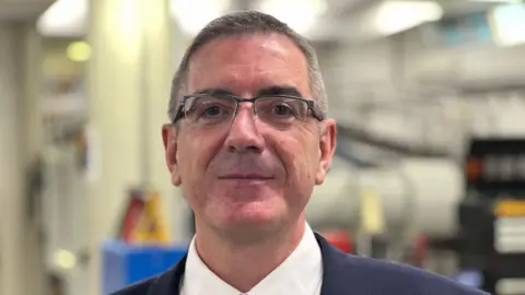A man wearing glasses and a suit with a white shirt is looking directly at the camera. He has grey hair and is cleanly shaven. He is standing indoors, with the background blurred.