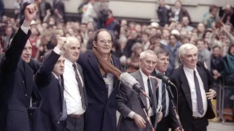 Reuters A photograph of seven men in suits standing in front of microphones, with a large crowd in the background. The two men on the right have raised their fists in the air in victory.