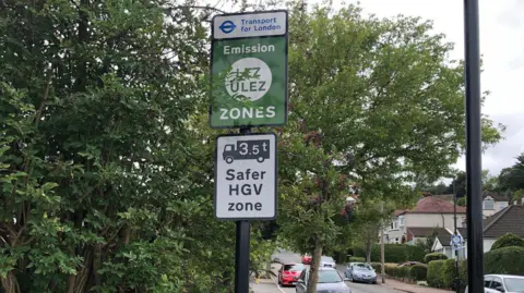 Jack Fiehn/BBC A TfL sign which shows drivers they are entering LEZ and ULEZ zones, there are trees visible in the background and a street with cars parked on it