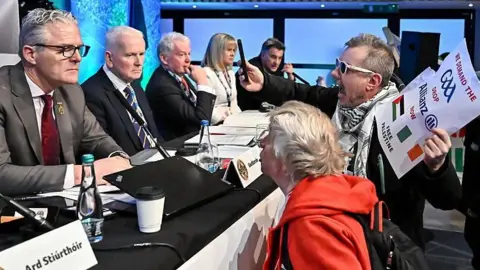 Getty Images Protestors talking to a panel while holding a sign and recording it with their phone.