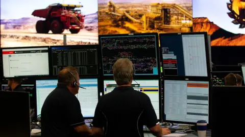 Rio Tinto Two men sit at a desk with dozens of trucks monitoring screens and other mining equipment