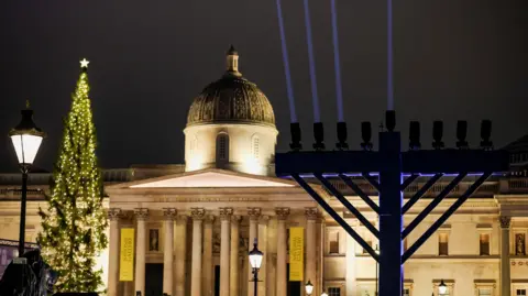 Reuters A menorah with light beams projecting from its branches stands in front of the National Gallery alongside a decorated Christmas tree during celebrations in Trafalgar Square in London on 16 December 2025.