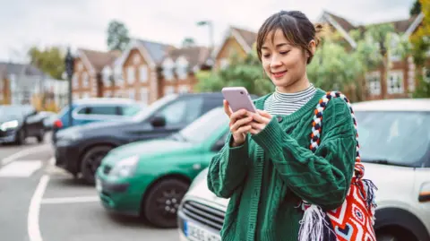 A young woman, dressed in a green cable-knit cardigan uses her phone in a car park, five cars are parked behind her, a silver, green, black, blue and black car. 