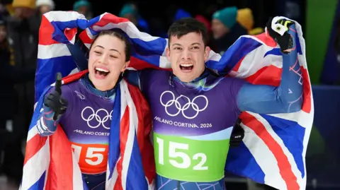 Great Britain's Matt Weston and Tabitha Stoecker, both wearing Team GB lycra kit and draped in a union flag, punch the air as they celebrate winning gold after the Skeleton Mixed Team Final at the Cortina Sliding Centre, on day nine of the Milano Cortina 2026 Winter Olympics, Italy.