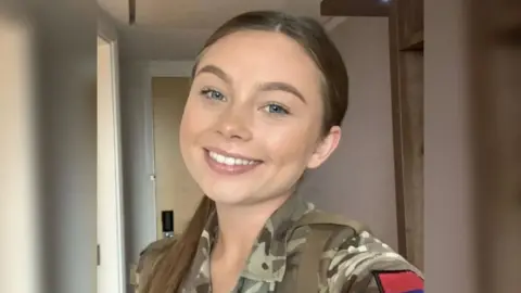 Family photo A selfie of Jaysley Beck wearing her military uniform, with her long brown hair slicked back into a low ponytail. She has blue eyes and is smiling at the camera.