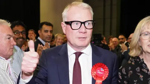 A man in a navy suit with a dark red tie and a red Labour rosette pinned to it. He has short, white hair and thick black glasses. He has his hand in the air and is giving a thumbs up, He is surrounded by a crowd of people