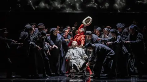 Johan Persson Performers in costume gather round a man lying in a chair on stage at the Bristol Hippodrome