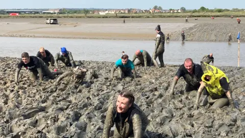 Men and women crawl through a muddy bank on a sunny day. They are covered in mud and some look exhausted as they make their way through. Other people can be seen behind them crawling through a flatter section of mud.