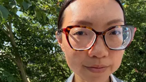 Provided Jenna Ho Marris, has large tortoise-shell glasses and dark hair tied up. There are trees in the background