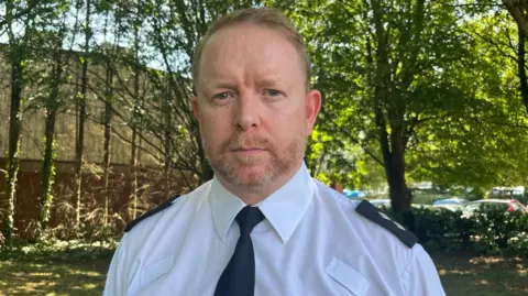Devon and Cornwall Police inspector Andy Wills stood in a white police shirt with a black tie. He is looking straight at the camera with a stern look on his face. He has a beard and is stood outside under some trees, with some buildings and cars in the background. 