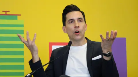 Russell Kane, a man wearing a white t-shirt and a black jacket is gesticulating with his hands as he talks into a microphone. He is pictured against a brightly coloured backdrop which is yellow and has additional block colour elements.