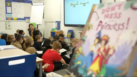 Jamie Niblock/BBC Children are sat on the floor of a classroom and drawing on whiteboards. Their teacher is kneeling while instructing them.