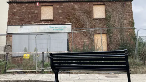 A black bench captured from behind at a lower angle. The view in front is off a derelict brick cottage with boarded up windows and metal fencing surrounding the front garden.