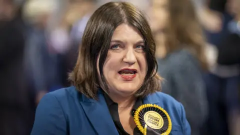 PA Media Susan Aitken, with dark hair and a blue jacket and yellow SNP rosette, in a medium close-up 