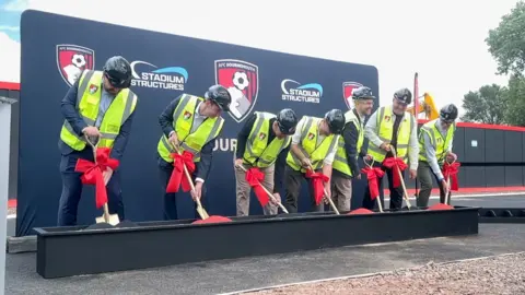 A group of seven men are all lined up wearing suits and yellow high vis vests. They are each holding a golden shovel with a red bow tied around the stems as they dig 'red' soil from a planter.