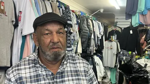 A man looking straight at the camera with a neutral expression. He is wearing a black cap and a black and white checked shirt and is standing in a clothes shop in front of a rail of T-shirts.