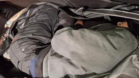 Home Office A man is pictured curled into a fetal position. He is in a large black holdall in the boot of a car. He is facing away from the camera. 