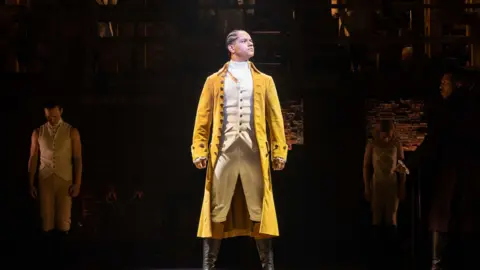 Danny Kaan A man stands spotlighted on stage in an 18th Century yellow frock coat, cream waistcoat and breeches. He is wearing long black boots and his head is turned to the side. He has braided black hair tied back. Behind him, other performers can just be seen in shadow.