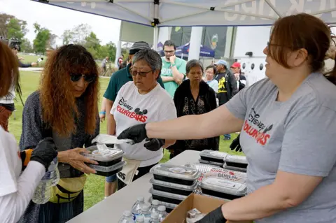 Women in T-shirts under a tent at a table, receive black boxes of food from woman wearing gray T-shirt and glasses