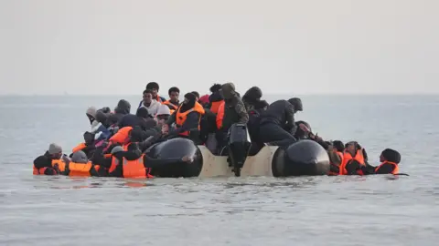 A small, crowded inflatable boat filled with people in bright orange life jackets departs from the beach at Gravelines, France. Several individuals are still in the shallow water, attempting to climb aboard.