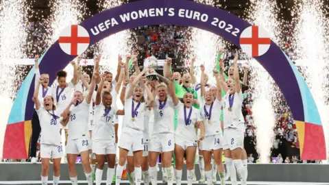 Getty Images England players in a white kit celebrate winning the UEFA Women's EURO 2022 Trophy after their sides victory during the UEFA Women's Euro 2022 final match between England and Germany at Wembley Stadium on July 31, 2022 in London, England. They all have medals which have a purple strap and gold medallion